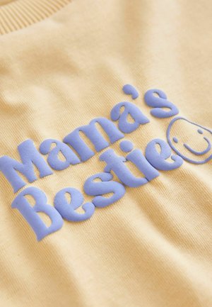Žltý pulóver s vystupujúcim modrým textom "Mama's Bestie" a úsmevom, vyrobený z mäkkej látky s rebrovaným golierom.
