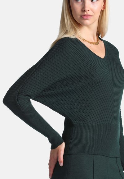 Femme aux cheveux blonds portant un pull côtelé vert foncé à col en V avec des manches dolman et un collier chaîne en or, main sur la hanche, regardant vers la droite.