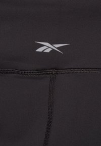 Svart atletiske leggings med en jevn tekstur, med et Reebok-logo i sølv og synlig søm langs sømmene.