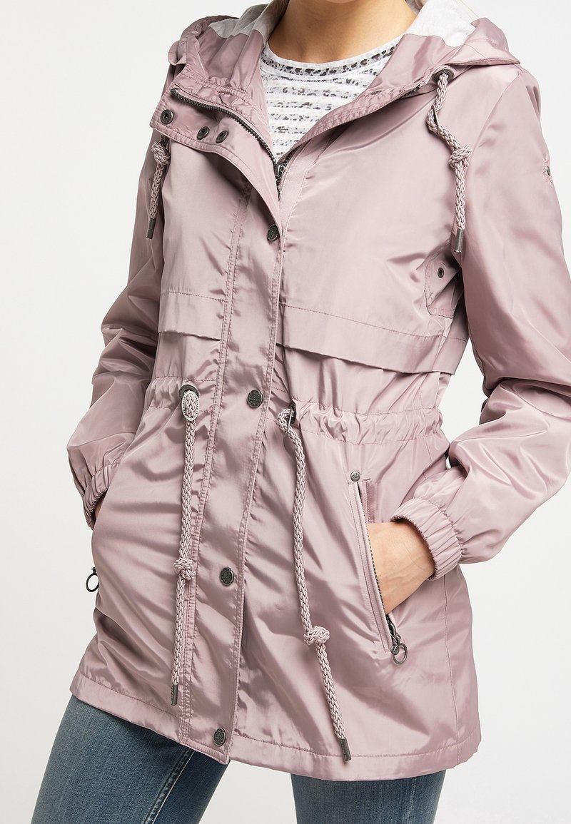 Femme portant une veste imperméable à capuche violet clair avec cordons, boutons sur le devant, poches zippées et poignets élastiques, sur une chemise rayée et un jean.