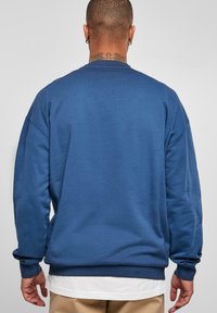 Felpa blu con scollo rotondo, spalle scese e polsini e orlo a coste. Tessuto liscio di un colore uniforme, senza motivi visibili.