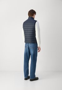 Granatowa kamizelka puchowa z poziomym pikowaniem, z wysokim kołnierzem, noszona na białej bluzie, zestawiona z niebieskimi dżinsami i czarnymi butami.