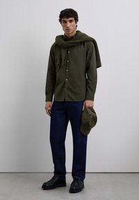 Young man wearing dark green button-up shirt and sweater around shoulders, dark blue pants, black shoes, holding a plaid cap in hand.