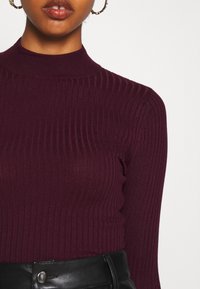 Pull côtelé à col montant de couleur bordeaux profond, doté de manches longues et d'une coupe ajustée, associé à un pantalon en cuir noir taille haute.