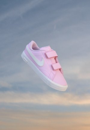 Baskets Nike rose clair avec semelle blanche et deux bandes velcro, présentées sur un fond de ciel doux et nuageux.
