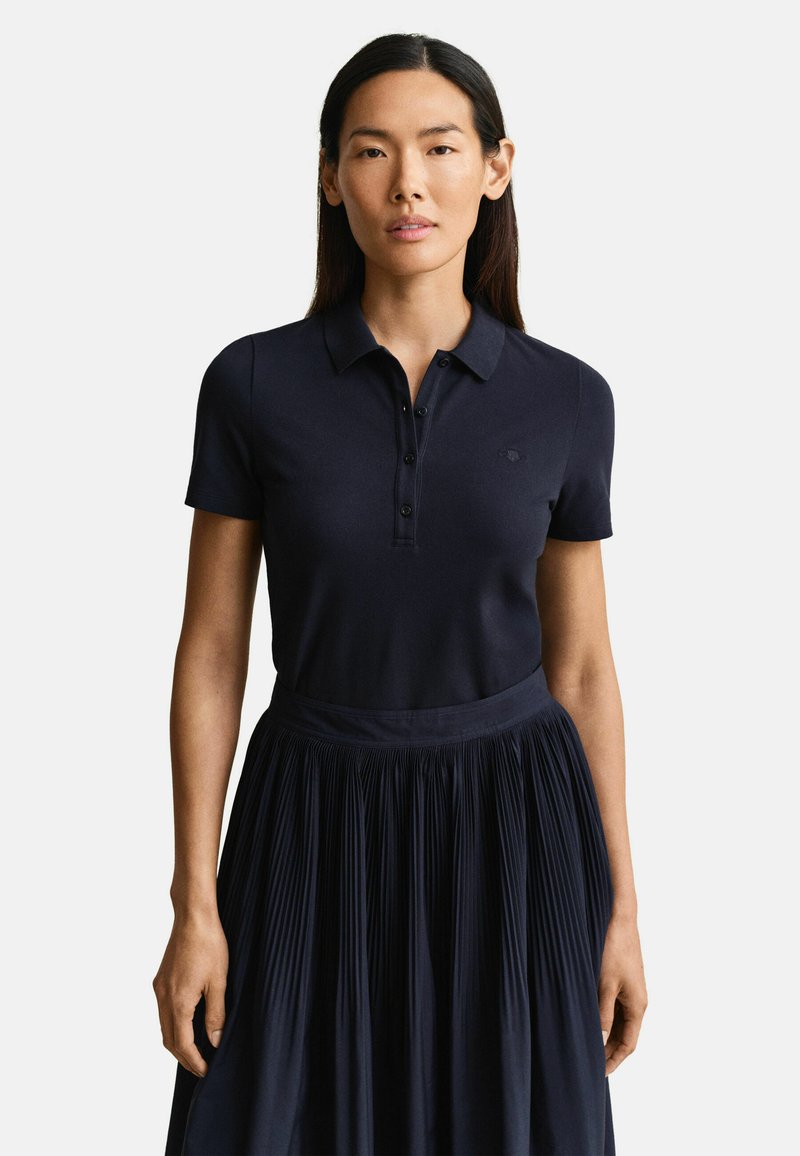 Femme aux longs cheveux bruns portant un polo bleu marine à manches courtes et une jupe plissée bleu marine, debout devant un fond uni.