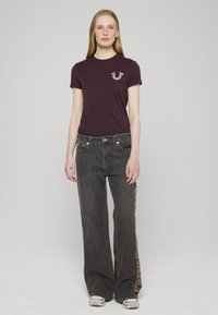 Camiseta de algodón color granate con un pequeño diseño gráfico en el pecho, combinada con jeans negros de pierna ancha que presentan bordado lateral y costuras de color claro.