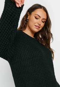 Svart stickad tröja med en strukturerad våffelmönster, vida ärmar och klassisk rund halsringning, som erbjuder en lös och avslappnad passform.