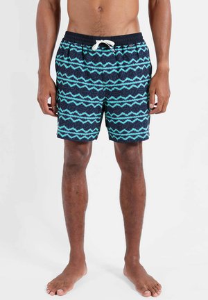 Torse et jambes masculins portant un short de bain bleu marine avec un motif zigzag turquoise et un cordon blanc, debout pieds nus sur fond blanc.