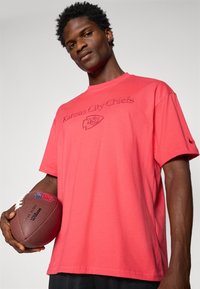 Rød kortærmet T-shirt med "Kansas City Chiefs" broderi og holdlogo. Modellen holder en brun fodbold med hvid syning.