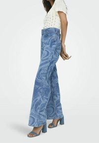 Top de punto blanco ajustado combinado con vaqueros de denim azul de pierna ancha con un patrón jaspeado y talle alto; usado con sandalias de tacón bloque azul.