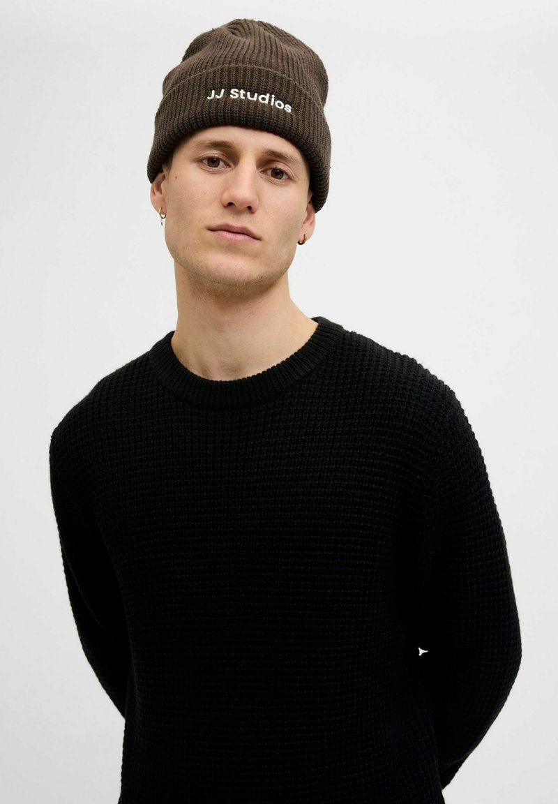 Bruine geribde beanie met het logo "JJ Studios" en een zwart gestructureerd gebreid sweater. Model staat met de armen langs het lichaam tegen een neutrale achtergrond.