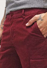 Bordové cargo kalhoty s hladkou texturou, přední kapsou a zapínáním na knoflík. Zobrazeny s rukou v kapse, což zvýrazňuje design pásku.