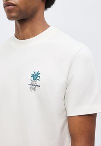 Κοντινό πλάνο ενός ατόμου που φοράει λευκό μπλουζάκι με μικρό μπλε και γκρι λογότυπο φυτού και κείμενο στο αριστερό στήθος.