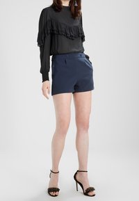 Žena v černé volánkové blůze s dlouhým rukávem, námořnických šortkách s vysokým pasem a knoflíky, a černých sandálech na vysokém podpatku s řemínky kolem kotníků.