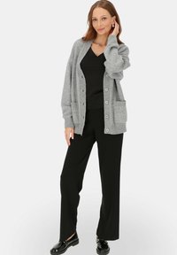 Szary dzianinowy kardigan z guzikiem z przodu, prążkowanymi mankietami i dwiema kieszeniami, w połączeniu z czarnymi spodniami i błyszczącymi czarnymi butami.