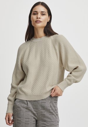 Beige gestrickter Sweatshirt mit strukturiertem Muster, geripptem Rundhalsausschnitt und Bündchen sowie überschnittenen Schultern. Kombiniert mit karierten Hosen.
