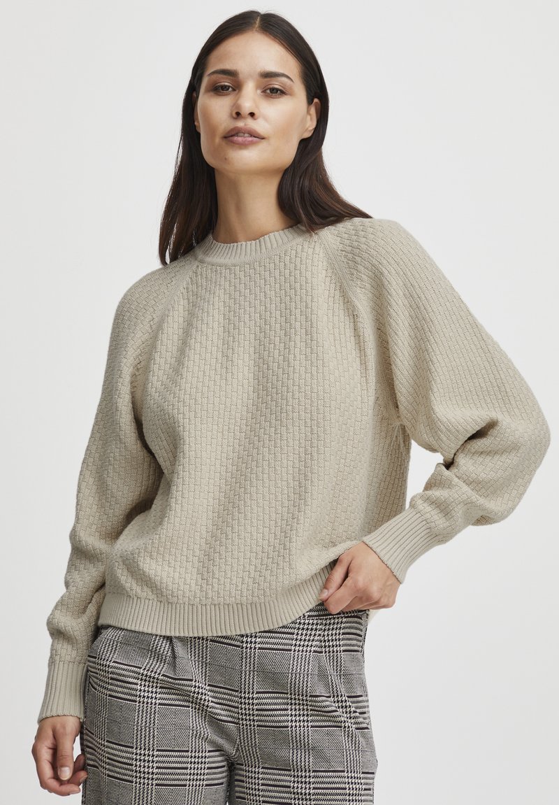 Pull en tricot beige avec un motif texturé, col rond côtelé et poignets, et épaules tombantes. Porté avec un pantalon à carreaux.
