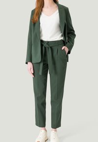Kobieta w ciemnozielonych spodniach z przewiązanym w pasie oraz dopasowanej marynarce, z białym topem w serek i jasnymi sneakersami, stoi z jedną ręką w kieszeni.