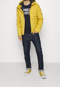 Muž v žluté prošívané bundě s kapucí, pod kterou má černé tričko s potiskem, tmavě modré zvlněné džíny a žluté plátěné tenisky, stojí s jednou rukou v kapse.