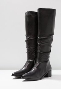 Botas de cuero negro hasta la rodilla con un diseño desaliñado, punta afilada y tacón cuadrado moderado. Textura suave con acentos de hardware mínimos.