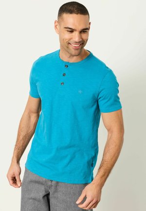 Bărbat zâmbitor purtând o cămașă henley cu mâneci scurte, de culoare albastru aprins, cu trei nasturi, asortată cu pantaloni gri deschis, privind în jos.