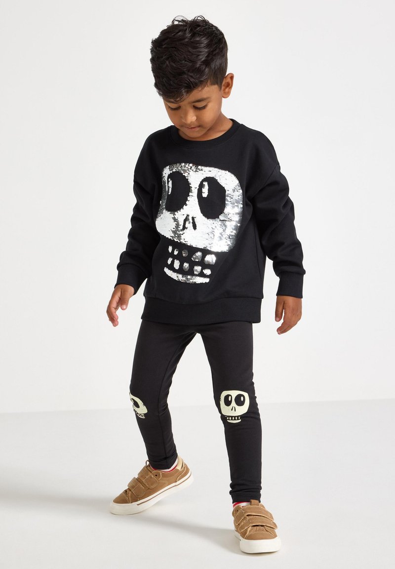 Sort sweatshirt med et sølvfarvet paliet skull-design, parret med sorte leggings med hvide skull-grafikker på knæene, og lyse sneakers.