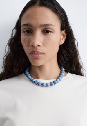 Kobieta o długich ciemnych włosach i piegach, ubrana w biały top i masywny niebieski naszyjnik z koralików na jednolitym tle.