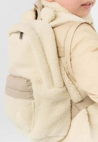 Kind draagt een beige jas en een crèmekleurige pluizige rugzak, bandjes zichtbaar, met gedeeltelijk zicht op gezicht en schouder.