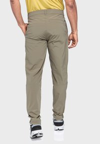 Helle olivfarbene Herren-Hosen aus weichem, flexiblem Material, im geraden Schnitt, mit seitlichen Taschen und einer Gesäßtasche, kombiniert mit schwarzen Sportschuhen.