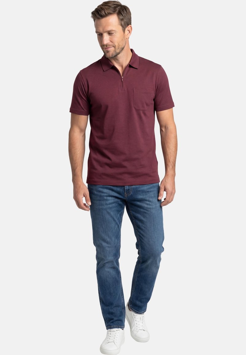 Homme portant un polo bordeaux à manches courtes avec fermeture éclair, jean bleu et baskets blanches, debout les mains détendues.