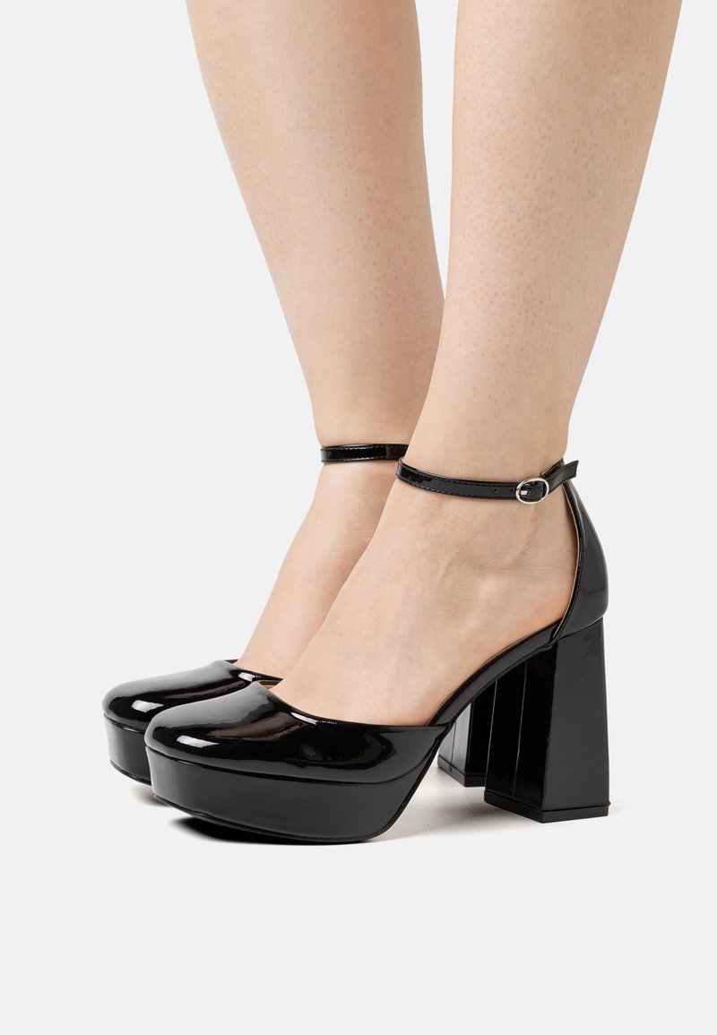 Talons compensés noirs brillants avec brides à la cheville portés pieds nus, avec des talons blocs épais et des bouts arrondis.