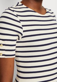 Randig ribbad topp i kräm- och marinblå färger. Har korta ärmar med guldknappdetaljer vid ärmkanterna.