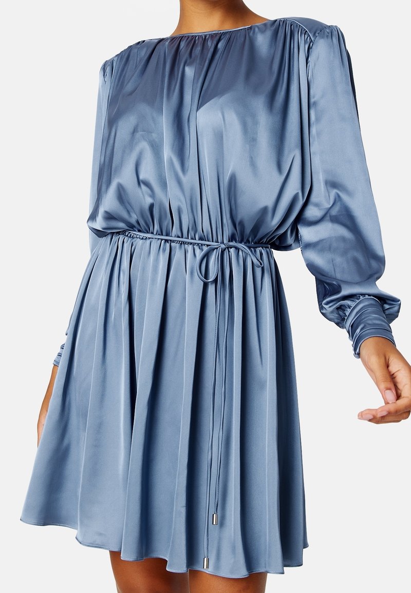Femme portant une robe bleue en satin à manches longues avec une taille cintrée par un lien et une jupe ample et fluide.