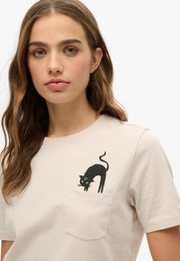 Camiseta de algodón beige con un gráfico de un gato negro en el bolsillo izquierdo. El diseño incluye una cola enroscada y detalles faciales distintivos.
