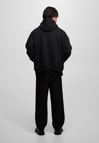 Sudadera negra de gran tamaño con capucha, hombros caídos y dobladillo de canalé, combinada con pantalones negros holgados y zapatos negros, vista desde atrás.