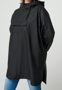 Sort vandtæt oversized jakke med forlommer, justerbar hætte og sideslidser; glat tekstur og afslappet pasform.