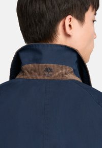 Chaqueta marina con un cuello de pana marrón; presenta detalles de costura y un logo en el cuello, fabricada con material suave.