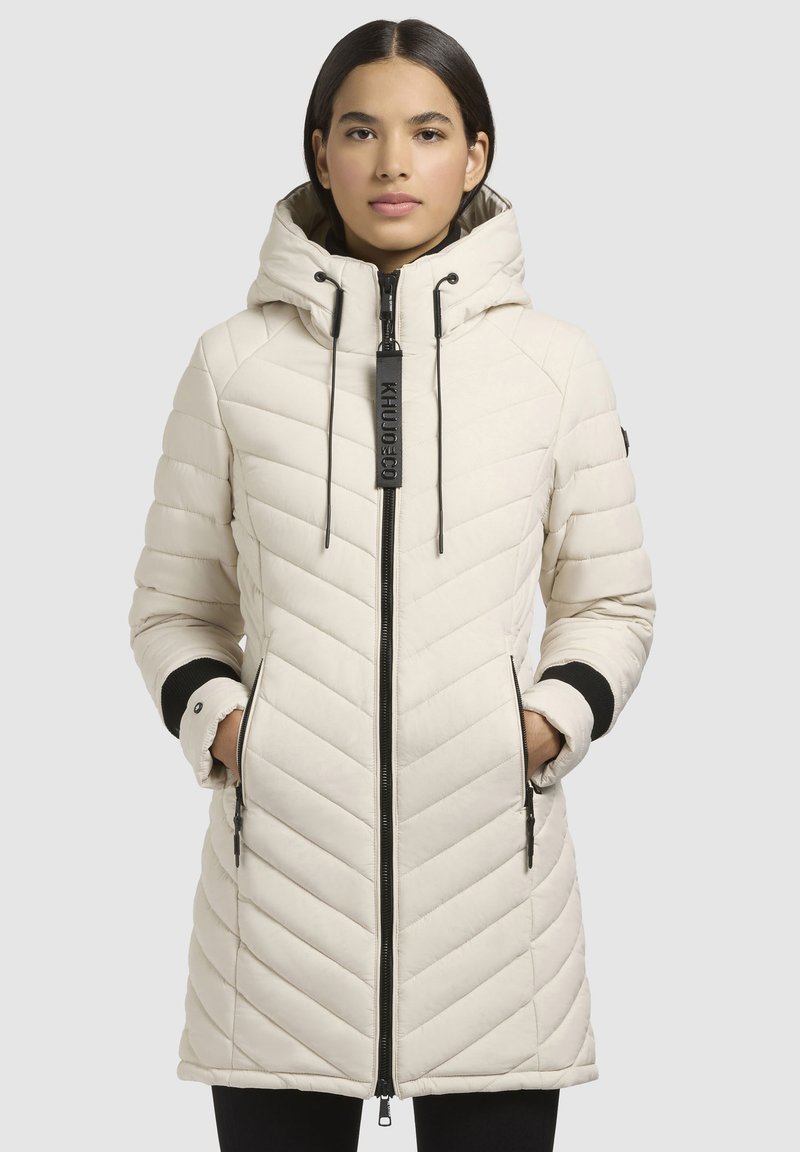 Lys beige pufjakke med chevron-quiltet design, hætte, sorte detaljer på manchetterne og lynlåslommer foran. Har en sort logo på lynlåsen.