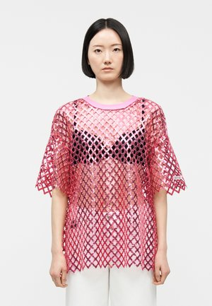 Žena s ravnom crnom kosom nosi široku, sjajnu ružičastu mrežastu bluzu s uzorkom rešetke preko crnog grudnjaka i bijelih hlača, stoji na bijeloj pozadini.