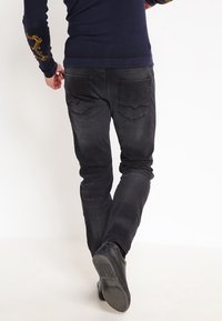 Vaqueros de mezclilla negros para hombre con un ajuste slim. Cuentan con bolsillos traseros con un diseño sutil. Combinados con un suéter azul marino.