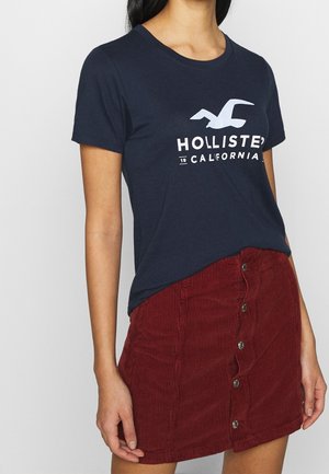Femme portant un t-shirt Hollister California bleu marine rentré dans une mini-jupe en velours côtelé rouge à boutons sur le devant, debout devant un fond uni.