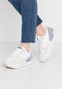 Sneakers bianchi con accenti in suede grigi e rosa, lacci testurizzati e una leggera suola a piattaforma, abbinati a jeans blu in denim arrotolati.