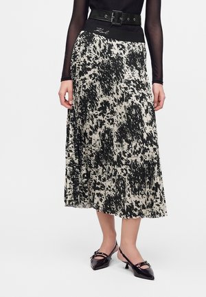 Femme portant une jupe midi plissée à motif abstrait en noir et blanc, ceinture noire, haut noir transparent à manches longues et chaussures noires à petits talons.