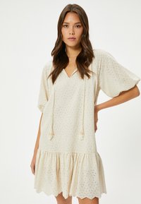 Cremefarbenes kurzes Kleid mit V-Ausschnitt, Puffärmeln und einem gestuften Rüschensaum. Verfügt über eine Lochmusterung und fransige Kordelzüge.
