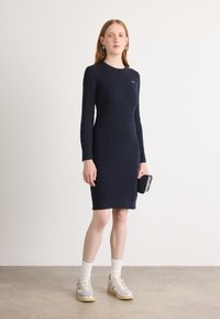 Jūrinės spalvos megzta suknelė ilgomis rankovėmis, su gijų raštu ir apvaliu iškirpimu, derinama su baltais kojiniais ir pilkais sportiniais bateliais.