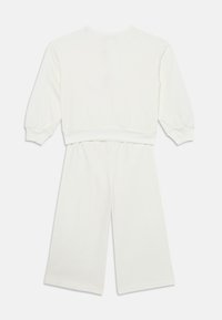 Weißes Zweiteiler-Outfit bestehend aus einem Langarmoberteil mit gesäumten Ärmeln und weit geschnittenen Hosen. Das Material wirkt weich und glatt mit minimalen Designelementen.