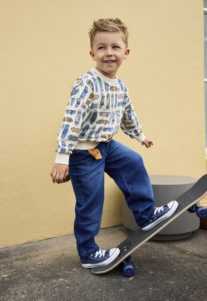 Young boy with skateboard-themed sweater and blue jeans steps onto skateboard near yellow wall outdoors.