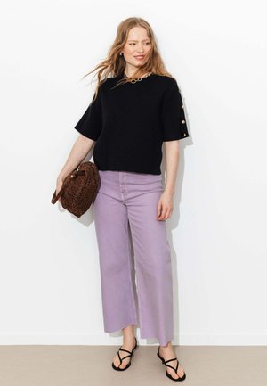 Femme aux cheveux roux ondulés porte un haut noir à manches courtes avec des boutons dorés, un pantalon large lavande, des sandales noires, et tient un sac à imprimé léopard.