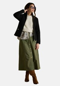 Čierna zipsová bunda, krémový prešívaný sveter, vzorovaná košeľa, olivovo zelená midi sukňa s gombíkmi a hnedé čižmy do kolien. Okrúhle okuliare.